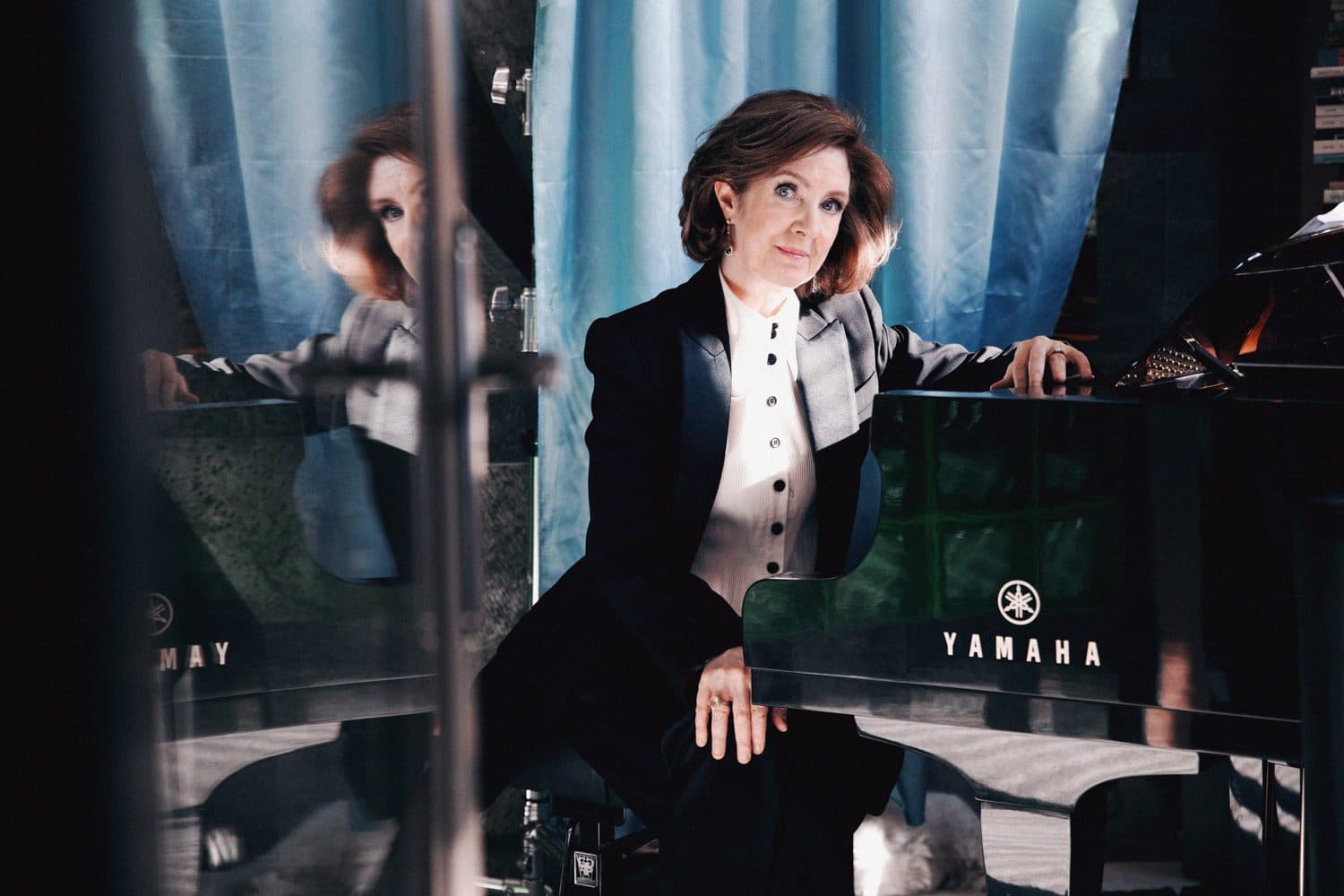 Woman in formal attire seated at a Yamaha grand piano with blue curtain background.