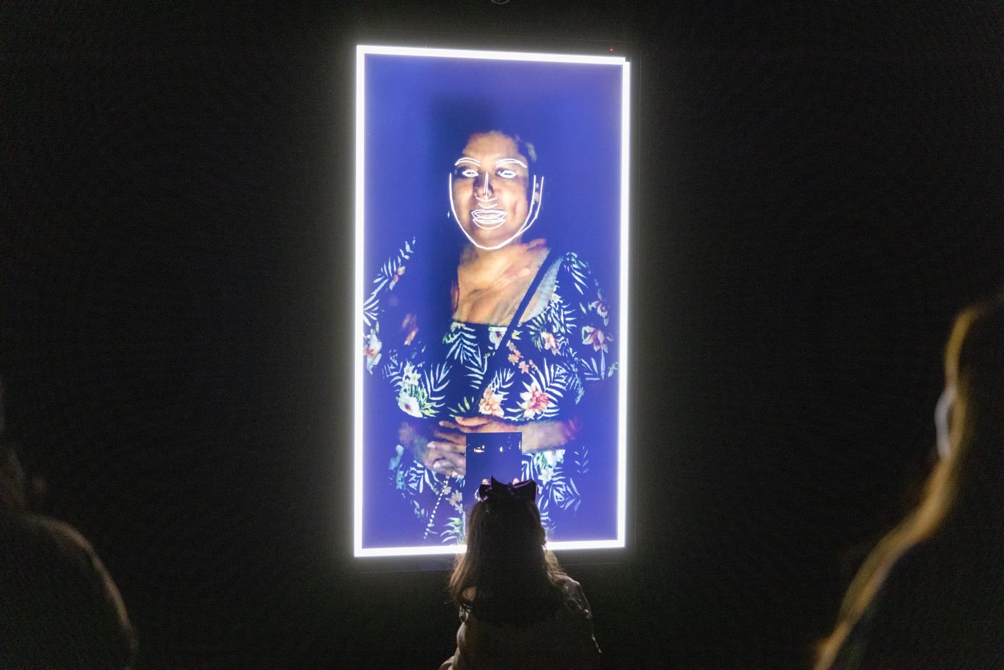A young girl stands facing a screen outside at night that displays a face blurring and combining into a composite portrait.